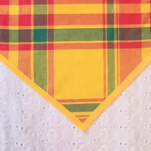 May include: A colorful, plaid table runner with a yellow border. The plaid pattern features red, orange, green, and brown squares and stripes. The runner is set against a white, eyelet fabric background.