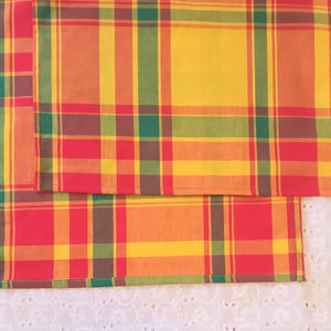 May include: Two red, yellow, orange, and green plaid tablecloths. The tablecloths are folded in half and are sitting on a white surface.