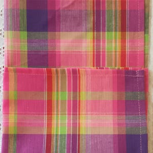 May include: Two pink and purple plaid fabric napkins. The napkins have a checkered pattern with pink, purple, green, orange, and brown stripes.