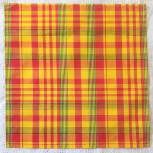 May include: A square, yellow, red, and green plaid fabric. The plaid pattern is made up of intersecting vertical and horizontal lines. The fabric is likely cotton or a similar material. The fabric is on a white lace surface.