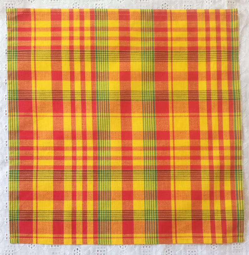 May include: A square, yellow, red, and green plaid fabric. The plaid pattern is made up of intersecting vertical and horizontal lines. The fabric is likely cotton or a similar material. The fabric is on a white lace surface.