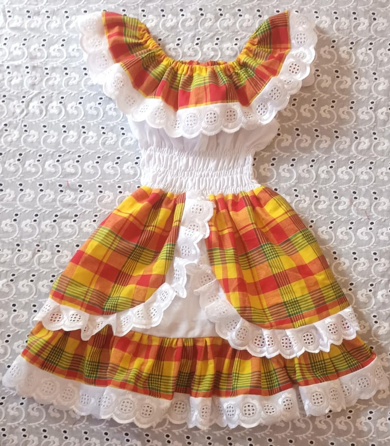 May include: A colorful plaid dress with a white eyelet trim. The dress features a ruffled neckline and skirt, with a fitted bodice. The plaid pattern includes yellow, red, and green hues. The dress is likely for a child.