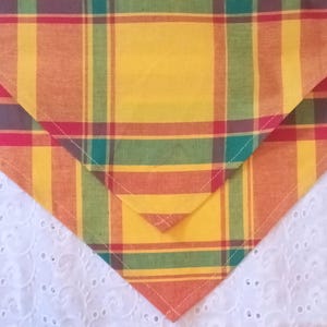 May include: A folded, plaid fabric in yellow, orange, red, green, and brown. The fabric is cut into a triangular shape, with the edges neatly sewn. The background is a white, eyelet fabric.