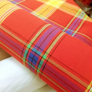 May include: Red fabric with a multi-colored plaid pattern. The fabric is folded and rolled up, showing the different colors and patterns.
