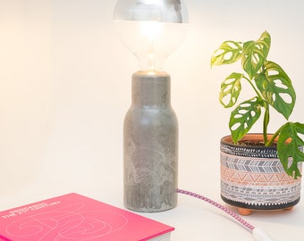 Puede incluir: Una lámpara de mesa de hormigón con una pantalla de vidrio transparente y una bombilla. La lámpara está enchufada y tiene un cable rosa y blanco. Una planta en maceta con hojas verdes está al lado de la lámpara. Un libro rosa con el título "Designing the 21st Century" está en primer plano.