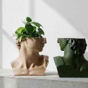 Op de afbeelding: Twee hoofd-vormige plantenbakken, één in een lichtbeige afwerking en de andere in een donker olijfgroene afwerking. Elke plantenbak heeft een klassiek buste-ontwerp met een hol bovengedeelte dat een kleine groene plant bevat.