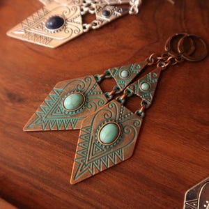 Pode incluir: Um par de brincos pendentes em tom de cobre com uma placa de metal em forma de diamante, apresentando padrões geométricos gravados e uma pedra oval de cor turquesa ao centro. Os brincos estão presos a argolas e incluem um pequeno detalhe metálico triangular.