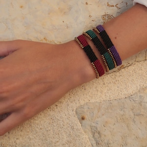 May include: Four colourful macrame bracelets with gold-coloured beads. The bracelets are in shades of red, black, green, and purple. The bracelets are worn on a wrist, with a white sleeve visible in the background.