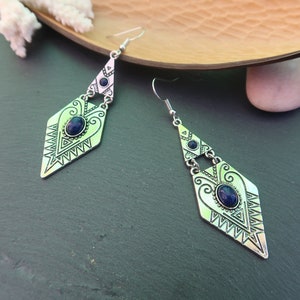Pode incluir: Um par de brincos pendentes em tom prateado com um pendente em forma de diamante, apresentando padrões geométricos gravados e uma pedra oval azul-escura central. Cada brinco inclui uma peça superior triangular com uma pedra azul a condizer.