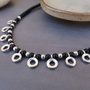 May include: A black beaded necklace with silver circle pendants. The necklace is on a gray surface.