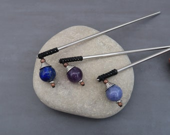 Puede incluir: Tres palitos de pelo plateados con envolturas de cordón negro y cuentas de piedras preciosas. Las cuentas son de lapislázuli, amatista y ágata azul.