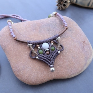 Op de afbeelding: Handgemaakte macramé ketting met een centrale hanger met een witte kraal, twee groene kralen en zilveren accenten. De ketting heeft een bruin koord en een lavendelkleurige kralenketting. De hanger is ongeveer 5 cm breed.