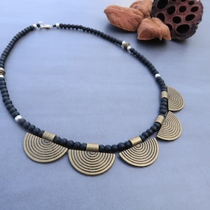 May include: A black beaded necklace with a silver clasp and five gold metal pendants. Each pendant is a half-circle with concentric rings.