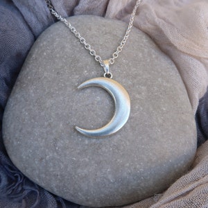 May include: A silver crescent moon pendant necklace on a silver chain. The necklace is displayed on a gray stone.