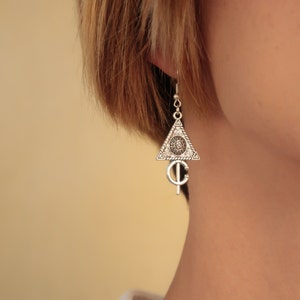 May include: Silver triangle earrings with a detailed design featuring a circle with a smaller circle inside and a symbol hanging below.