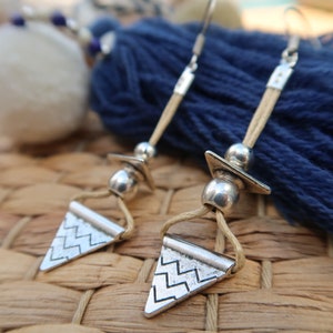 Puede incluir: Pendientes plateados con un colgante triangular con un diseño en zigzag. Los pendientes tienen un cordón beige y detalles plateados, con una borla azul oscuro en el fondo. Los pendientes miden aproximadamente 5 cm de largo.