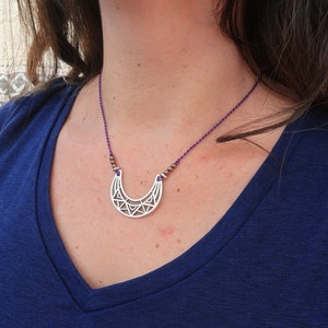 May include: A silver crescent moon necklace with a geometric design. The necklace is on a purple cord with small brown beads.