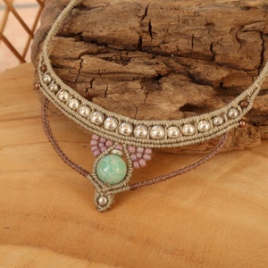 May include: A beige macrame necklace with silver beads and a green stone pendant. The necklace is on a wooden surface.