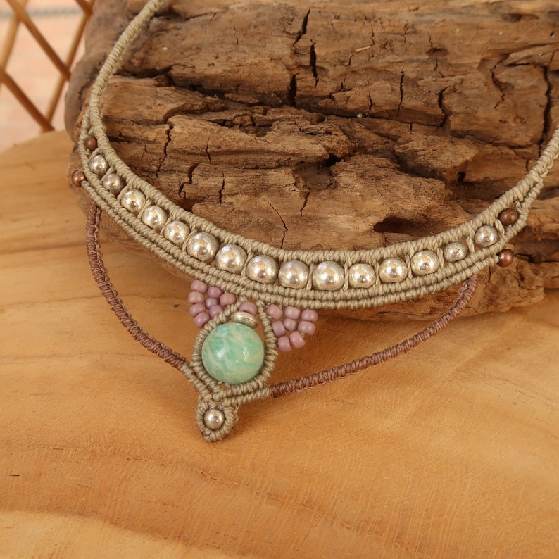 May include: A beige macrame necklace with silver beads and a green stone pendant. The necklace is on a wooden surface.