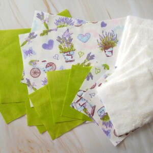 Op de afbeelding: Een set van stoffen vierkanten in verschillende kleuren en patronen. De set bevat groene vierkanten, witte vierkanten met een lavendelprint en witte badstof vierkanten.