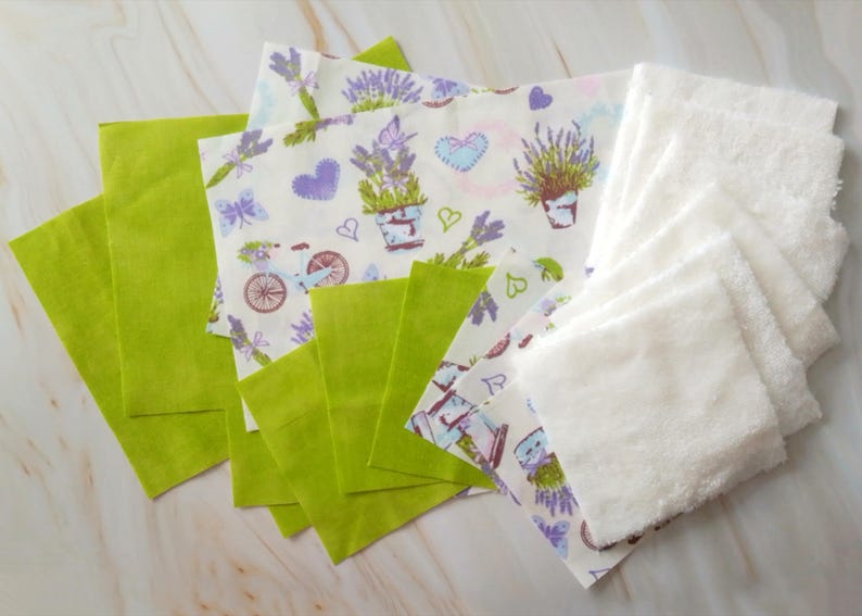 Op de afbeelding: Een set van stoffen vierkanten in verschillende kleuren en patronen. De set bevat groene vierkanten, witte vierkanten met een lavendelprint en witte badstof vierkanten.
