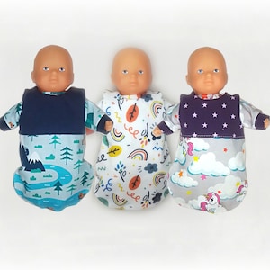 Peut inclure: Trois poupées bébés portant des gigoteuses différentes. Les gigoteuses sont en tissu doux et ont des motifs différents. Une gigoteuse a un motif de montagne et de forêt bleu et vert, une autre a un motif blanc et arc-en-ciel, et la dernière a un motif gris et licorne.