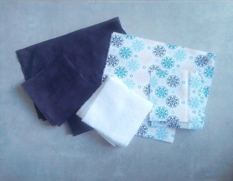 Op de afbeelding: Een set stoffen vierkanten in verschillende kleuren en patronen. De set bevat een donkerblauw vierkant, een wit vierkant en een gepatroneerd vierkant met blauwe en witte bloemen.