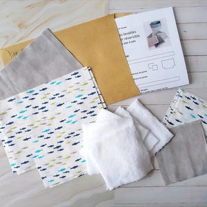 Op de afbeelding: Een set stoffen restjes voor naaiprojecten. De restjes zijn in verschillende grijstinten en wit met een blauw en groen vispatroon. De restjes zijn gerangschikt op een licht houten oppervlak met een vel papier met instructies voor het maken van omkeerbare, wasbare doeken.