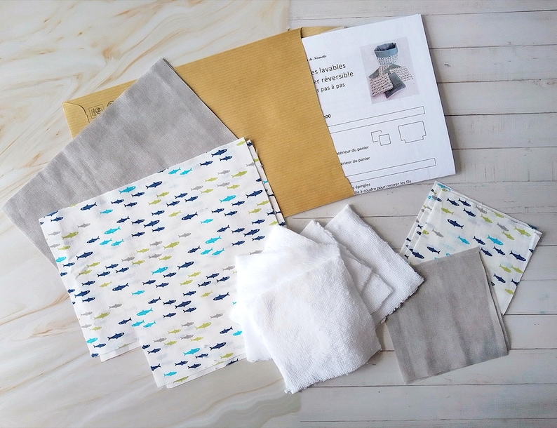 Op de afbeelding: Een set stoffen restjes voor naaiprojecten. De restjes zijn in verschillende grijstinten en wit met een blauw en groen vispatroon. De restjes zijn gerangschikt op een licht houten oppervlak met een vel papier met instructies voor het maken van omkeerbare, wasbare doeken.