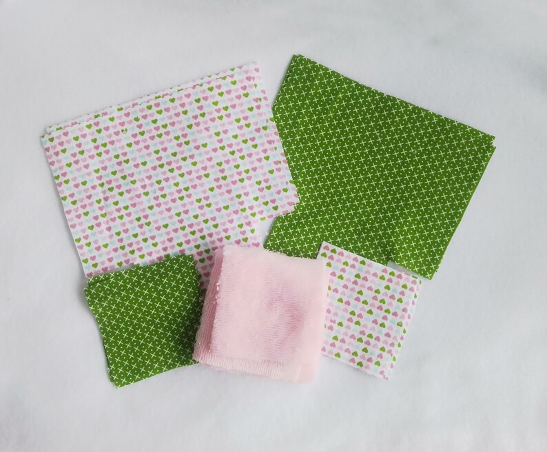 Op de afbeelding: Assortiment van stoffen vierkanten in roze, groen en wit met verschillende patronen, waaronder harten en een geometrisch ontwerp. Een roze washandje is ook inbegrepen.