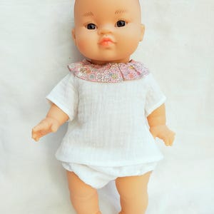 Peut inclure: Une poupée bébé à la peau légèrement brune, portant une chemise blanche avec un col fleuri rose et une culotte blanche. La poupée a les yeux noirs et une expression neutre.