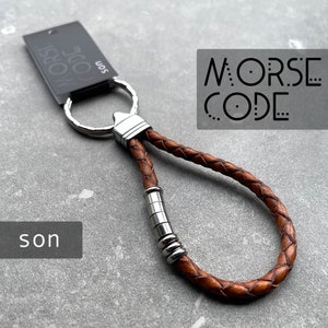May include: Brown leather keychain with a silver key ring and a silver metal bead accent. The keychain has the words "MORSE CODE" printed on it.