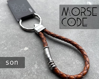 Puede incluir: Llavero de cuero marrón con un anillo de llave plateado y un acento de cuentas de metal plateado. El llavero tiene las palabras "MORSE CODE" impresas.