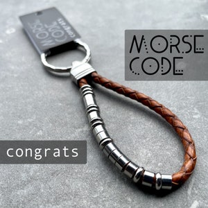 May include: A brown leather keychain with a silver ring and a silver chain of beads. The keychain has the words "MORSE CODE" printed on it. The text "congrats" is also visible in the image.