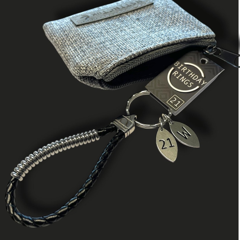 May include: A black leather keychain with a silver accent and a silver key ring. The keychain has a tag that says "BIRTHDAY RINGS" and two silver charms with the number "21" and the letter "W". A gray fabric pouch is in the background.