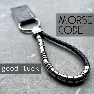 May include: A black leather keychain with silver metal detailing and a metal ring. The keychain features the words "MORSE CODE" and "good luck".