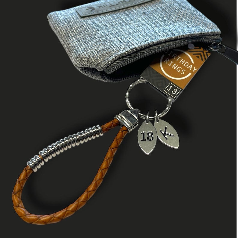 May include: A brown leather keychain with a silver accent and a braided design. The keychain has two silver leaf-shaped charms with the number 18 and a letter. A gray fabric pouch is in the background.