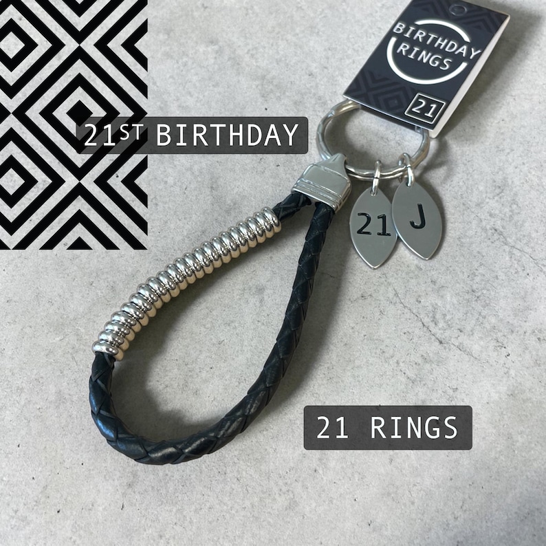 May include: A black leather keychain with silver accents and two oval charms. The charms are engraved with the number "21" and the letter "J". The keychain is labeled "21st Birthday" and "21 Rings".