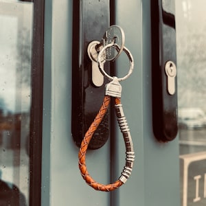 K&ouml;nnte beinhalten: Ein Schl&uuml;sselanh&auml;nger aus braunem geflochtenem Leder mit einem silberfarbenen Metallring und einer Reihe kleiner, zylindrischer silberfarbener Metallperlen an einer Seite der Schlaufe. Der Anh&auml;nger steckt an einem Schl&uuml;ssel im T&uuml;rschloss.