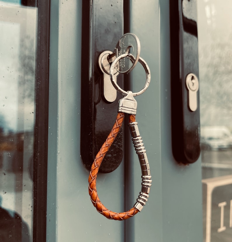 K&ouml;nnte beinhalten: Ein Schl&uuml;sselanh&auml;nger aus braunem geflochtenem Leder mit einem silberfarbenen Metallring und einer Reihe kleiner, zylindrischer silberfarbener Metallperlen an einer Seite der Schlaufe. Der Anh&auml;nger steckt an einem Schl&uuml;ssel im T&uuml;rschloss.