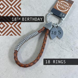 May include: A brown leather keychain with silver accents and two oval charms. The charms are engraved with "18 S" and the tag reads "BIRTHDAY RINGS 18". The text "18TH BIRTHDAY" and "18 RINGS" are also visible.