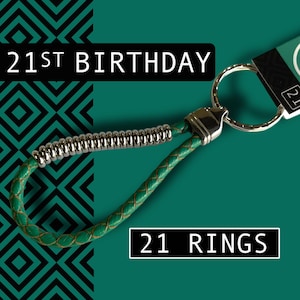 May include: Green leather keychain with a silver metal accent and a large metal ring. The text "21ST BIRTHDAY" and "21 RINGS" are displayed on the image. The background is teal with a black geometric pattern.