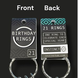 May include: A set of 21 rings on a keychain, with a black and white tag. The front of the tag reads "BIRTHDAY RINGS" and "21". The back of the tag says "21 RINGS" and "ONE RING TO CELEBRATE EACH SPECIAL YEAR".