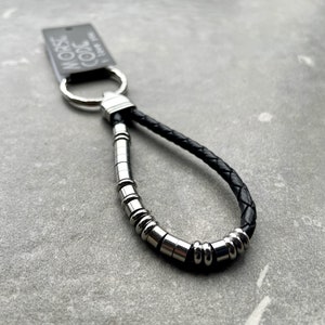 K&ouml;nnte beinhalten: Ein Schl&uuml;sselanh&auml;nger aus schwarzem geflochtenem Leder mit einem silberfarbenen Metallring und einer Reihe von zylindrischen und kugelf&ouml;rmigen silberfarbenen Perlen in einem Muster. Ein rechteckiges Etikett am Ring zeigt den Text MORSE CODE I love you.