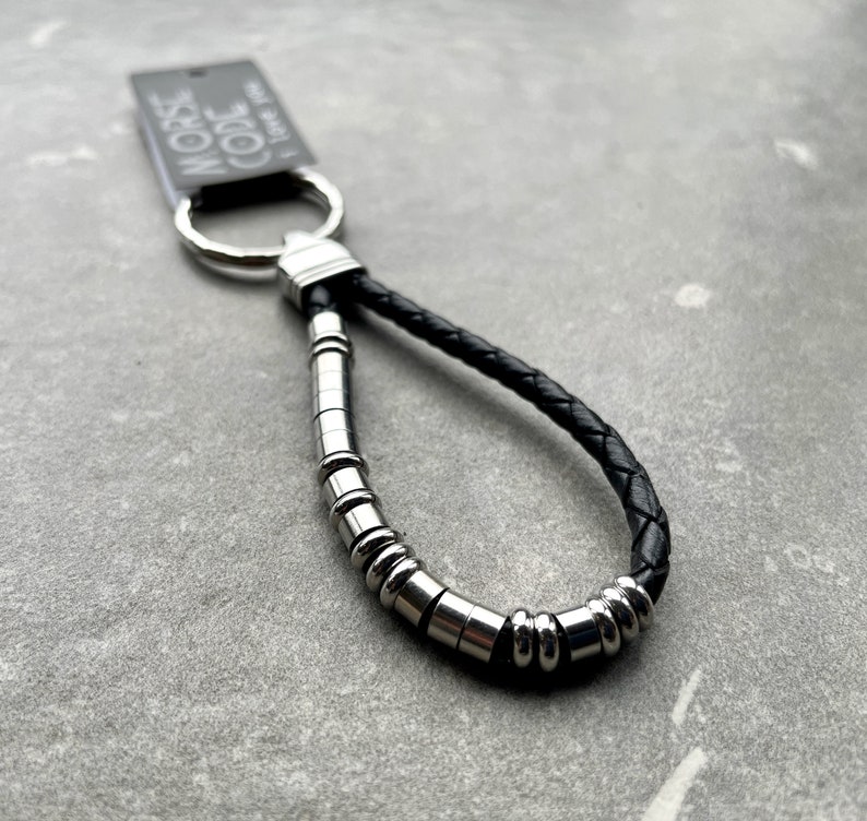 K&ouml;nnte beinhalten: Ein Schl&uuml;sselanh&auml;nger aus schwarzem geflochtenem Leder mit einem silberfarbenen Metallring und einer Reihe von zylindrischen und kugelf&ouml;rmigen silberfarbenen Perlen in einem Muster. Ein rechteckiges Etikett am Ring zeigt den Text MORSE CODE I love you.