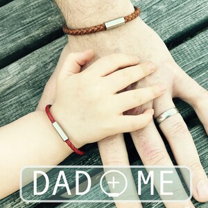 Puede incluir: Dos personas llevando pulseras de cuero a juego con detalles plateados. Las pulseras son trenzadas y tienen un dise&ntilde;o sencillo. El texto "DAD + ME" es visible en la imagen.