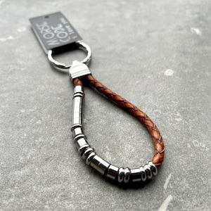 K&ouml;nnte beinhalten: Ein Schl&uuml;sselanh&auml;nger aus braunem geflochtenem Leder mit einer Reihe silberfarbener zylindrischer und ringf&ouml;rmiger Metallperlen auf einer Seite. Ein silberner Metallring ist oben befestigt und h&auml;lt ein schwarzes Etikett mit der Aufschrift MORSE CODE I love you.