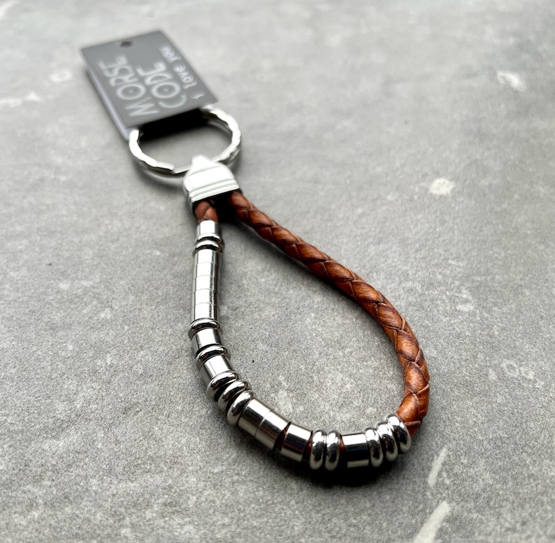 K&ouml;nnte beinhalten: Ein Schl&uuml;sselanh&auml;nger aus braunem geflochtenem Leder mit einer Reihe silberfarbener zylindrischer und ringf&ouml;rmiger Metallperlen auf einer Seite. Ein silberner Metallring ist oben befestigt und h&auml;lt ein schwarzes Etikett mit der Aufschrift MORSE CODE I love you.