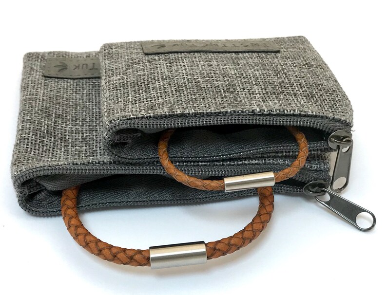 Puede incluir: Dos estuches grises con cremallera con un brazalete de cuero marr&oacute;n en su interior. Los estuches est&aacute;n hechos de una tela texturizada y uno de ellos tiene la palabra "TUK" impresa.