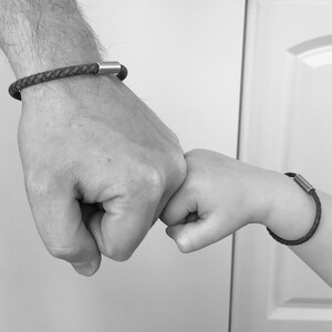 Puede incluir: Dos personas, una con una pulsera de cuero negro con un cierre plateado, se dan un pu&ntilde;etazo. La pulsera en la mu&ntilde;eca m&aacute;s peque&ntilde;a tiene un cierre plateado m&aacute;s peque&ntilde;o.